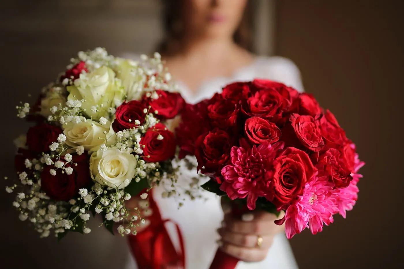 Deux bouquets de mariage tenus par une mariée vêtue de blanc. Le premier bouquet est composé de roses blanches et de gypsophile, symbolisant la pureté, tandis que le second est un arrangement audacieux de roses rouges et roses vifs, représentant la passion. Les bouquets sont tenus devant une robe de mariée blanche, capturant l'essence de l'amour et de l'engagement.