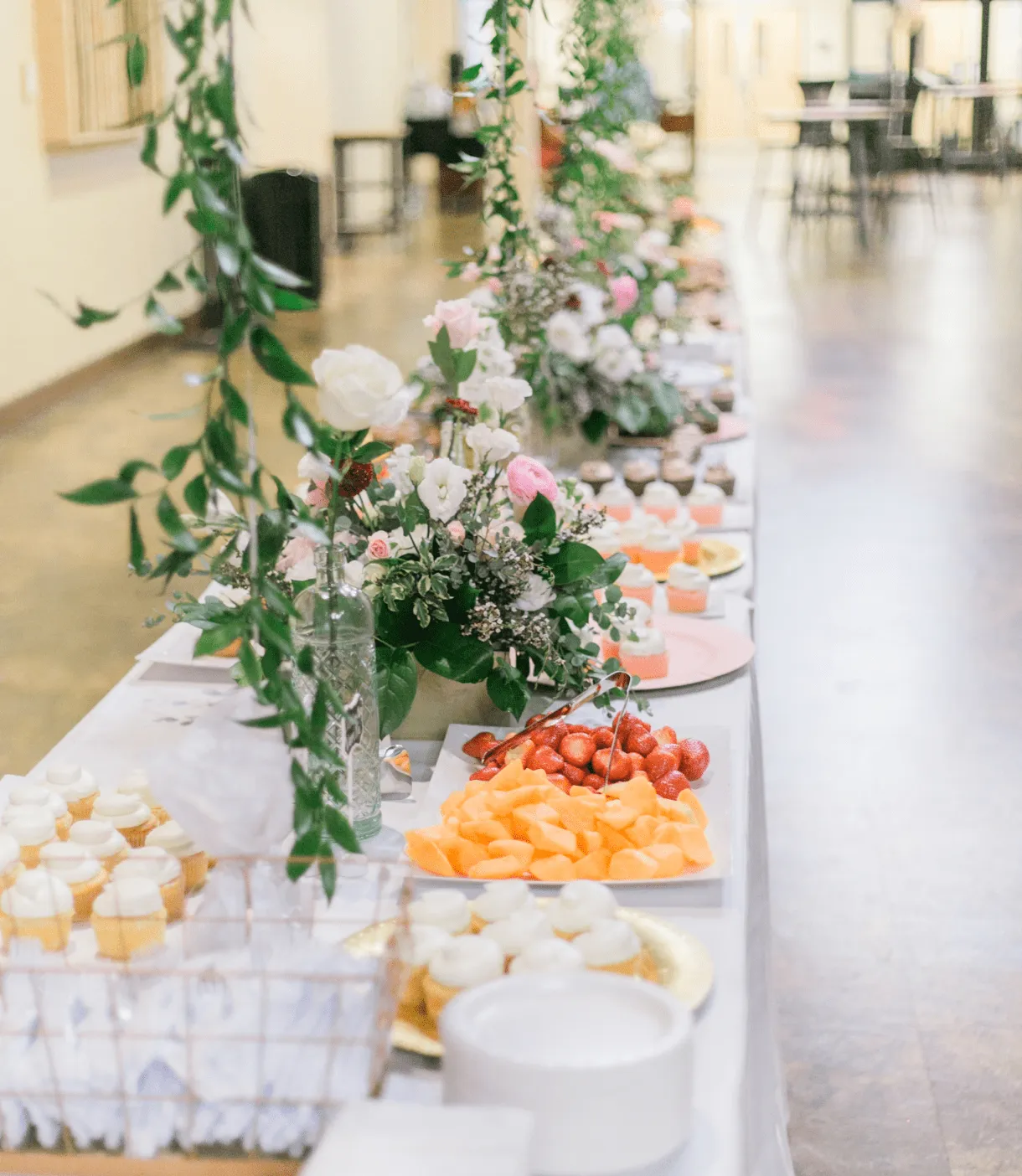 Une table de réception de mariage élégamment décorée avec des arrangements floraux délicats en blanc et rose, entourés de verdure. La table présente différents plateaux de fruits et de desserts - tranches d'oranges, fraises, et petits gâteaux blancs - créant une ambiance raffinée et invitante pour un événement privé.