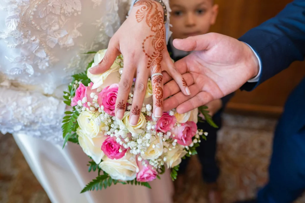 Gros plan sur les mains d'un couple lors d'une cérémonie de mariage. La main de la mariée, ornée de délicats motifs de henné traditionnels, tient un bouquet de roses blanches et roses, avec des gypsophiles blanches. La main du marié, vêtu d'un costume bleu, repose à côté. L'image capture l'intimité et la beauté d'un moment sacré, mêlant tradition culturelle et amour.