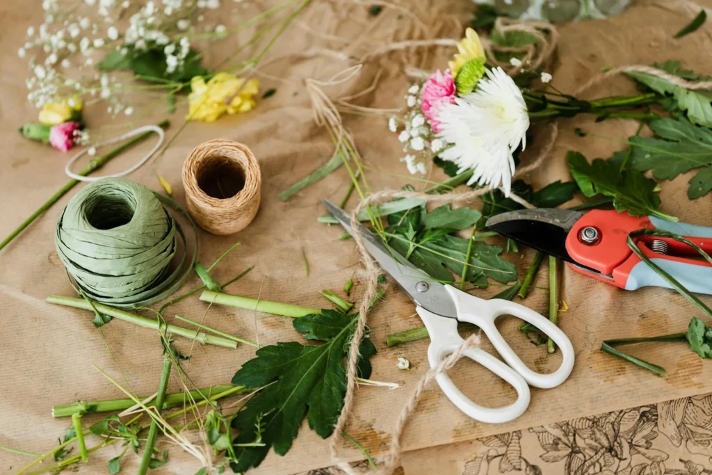 Un arrangement de matériaux de composition florale sur un fond de papier kraft : des bobines de ficelle verte et naturelle, des ciseaux blancs, des ciseaux de taille-haie rouges, des fleurs blanches et colorées, des feuilles vertes et des brins d'herbe. L'image capture l'essence de l'art floral, avec ses outils et ses matériaux organiques.